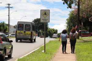 #pracegover a imagem moctra duas mulheres caminhando na calcada ao lado de uma plca de advertencia quanto a travessia de escolares, enquanto uma van de transporte escolar circula na via